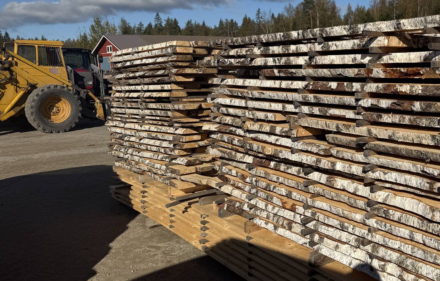 A pallet of curly birch boards stacked and strapped in the yard, a wheel loader and a red farm building behind